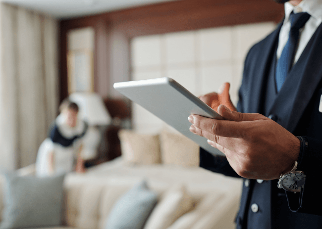 Hotel manager using a tablet to monitor operational implementation while housekeeping staff prepare a guest room